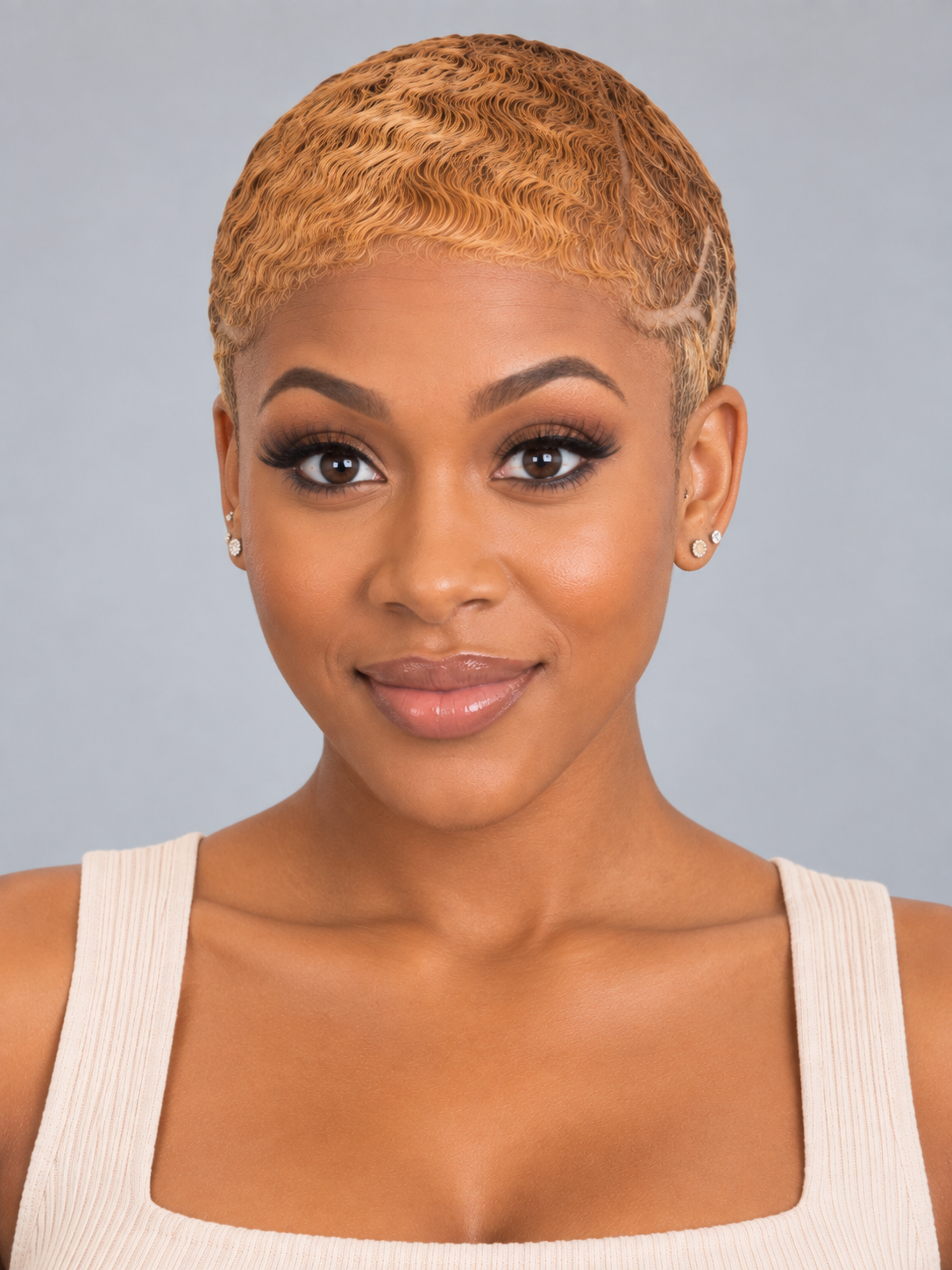 A woman with a short, textured blonde pixie cut wearing a cream ribbed tank top, photographed against a solid gray studio background.