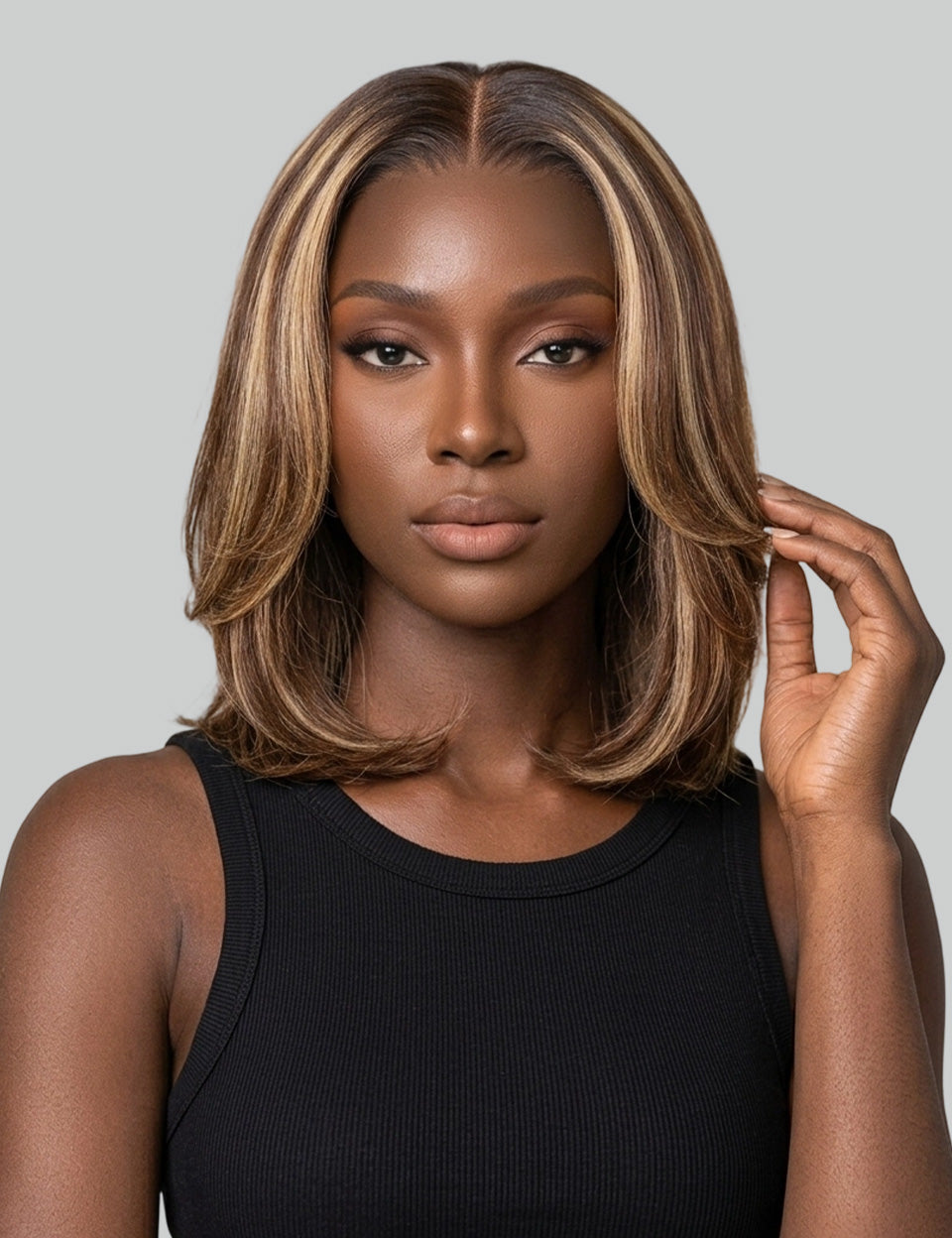 A woman modeling a shoulder-length layered wig with side-swept bangs, featuring warm brown and blonde highlights, against a solid gray background.