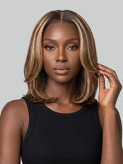 A woman modeling a shoulder-length layered wig with side-swept bangs, featuring warm brown and blonde highlights, against a solid gray background.