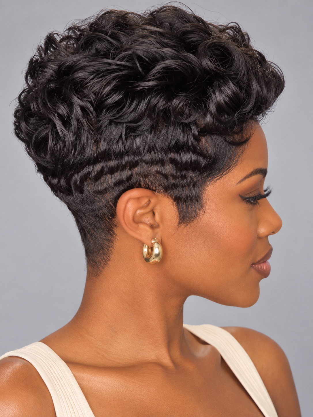 A woman shown in side profile wearing a short, curly black wig with a high-impact fade and gold hoop earrings against a solid gray background.