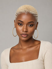 A close-up portrait of a model with short blonde hair wearing large gold hoop earrings and a cream ribbed top against a neutral gray background.