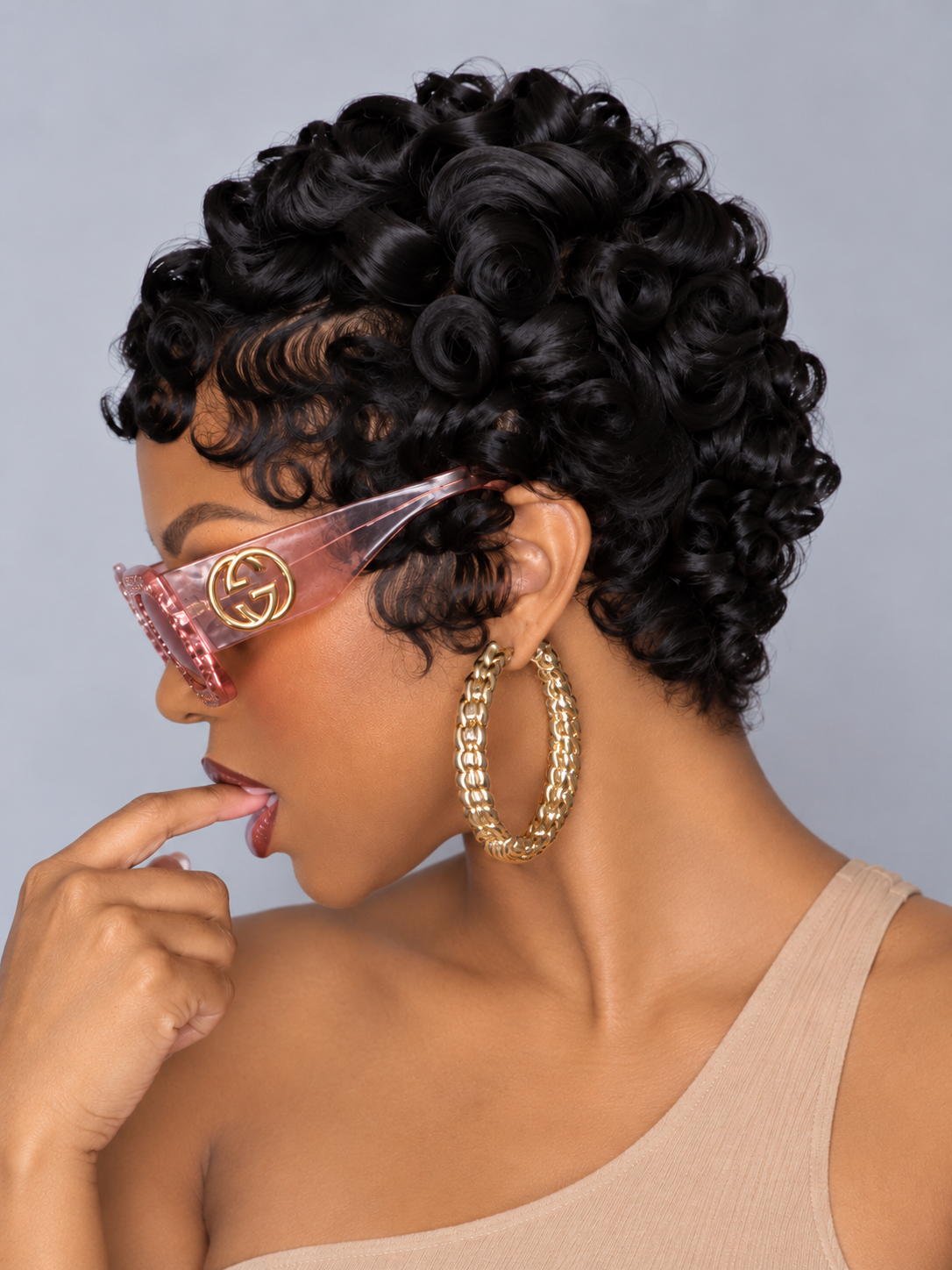 A woman with short, glossy, dark curly hair is shown in profile against a light gray studio background. She is wearing pink translucent sunglasses with a gold interlocking G logo, large gold hoop earrings, and a beige ribbed top.