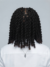Back view of a person with long, dark brown to black coiled dreadlocks or twists, wearing a white shirt and a silver cross necklace against a light gray background.