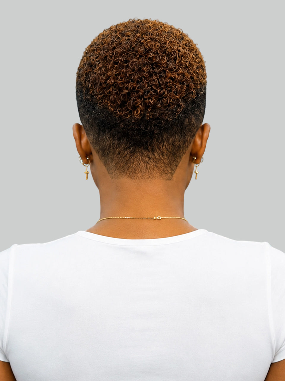 A woman in profile with a short, curly red hairstyle and shaved side details, wearing gold hoop earrings and a cream halter top against a light gray background.