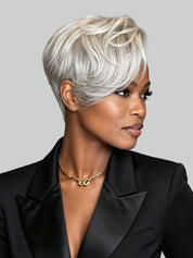 A woman with a short, layered silver-gray wig styled with soft waves, wearing a black blazer and gold necklace against a solid gray background.