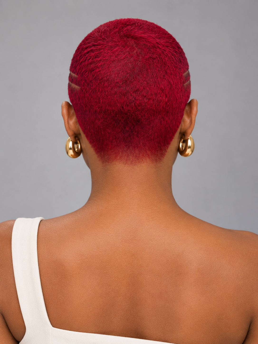 Back view of a person with a short, vibrant magenta-red pixie cut, wearing gold hoop earrings and a white top against a solid gray background.