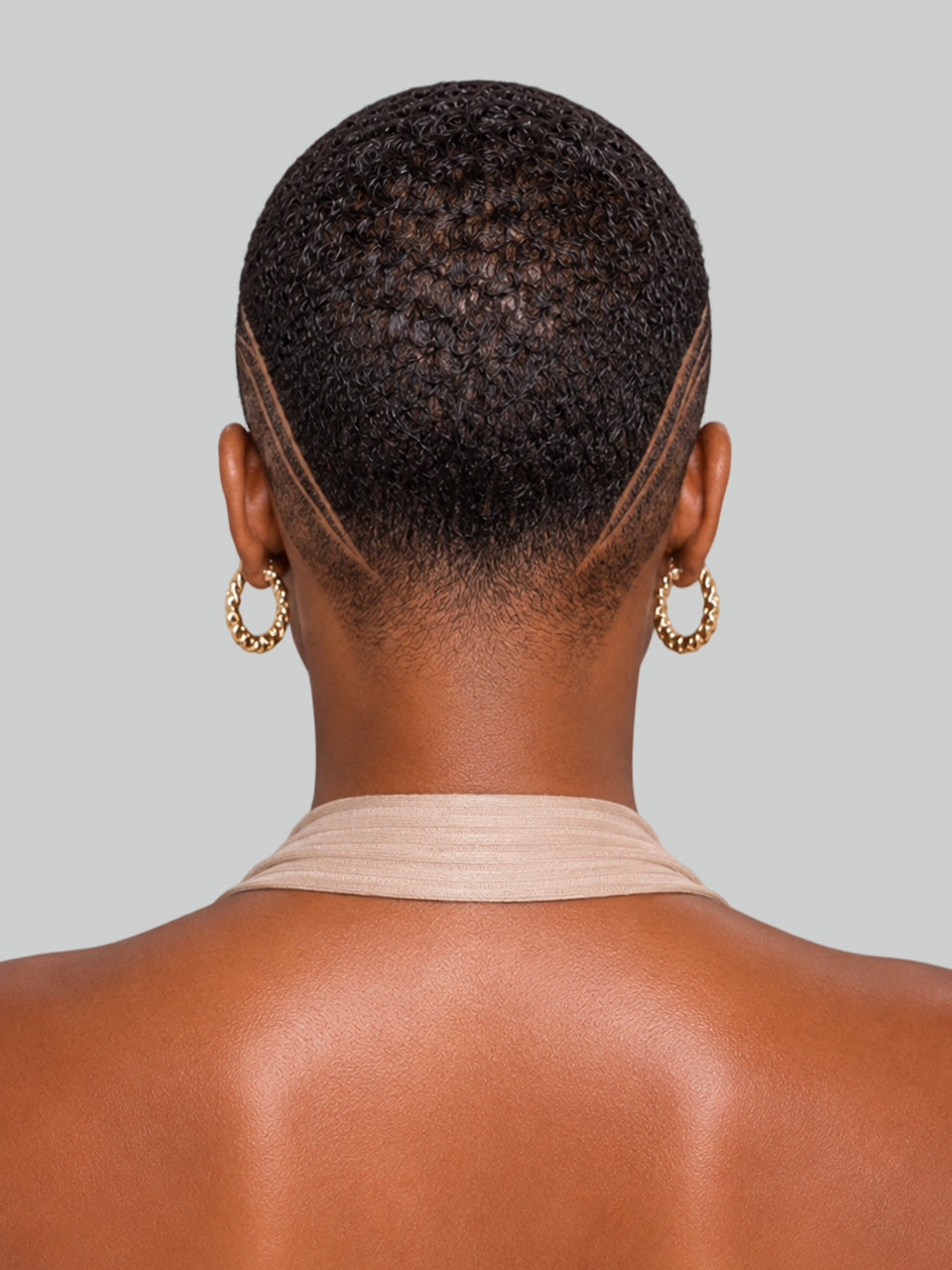 Rear view of a person with short textured hair wearing gold hoop earrings and a beige halter top against a light gray background.