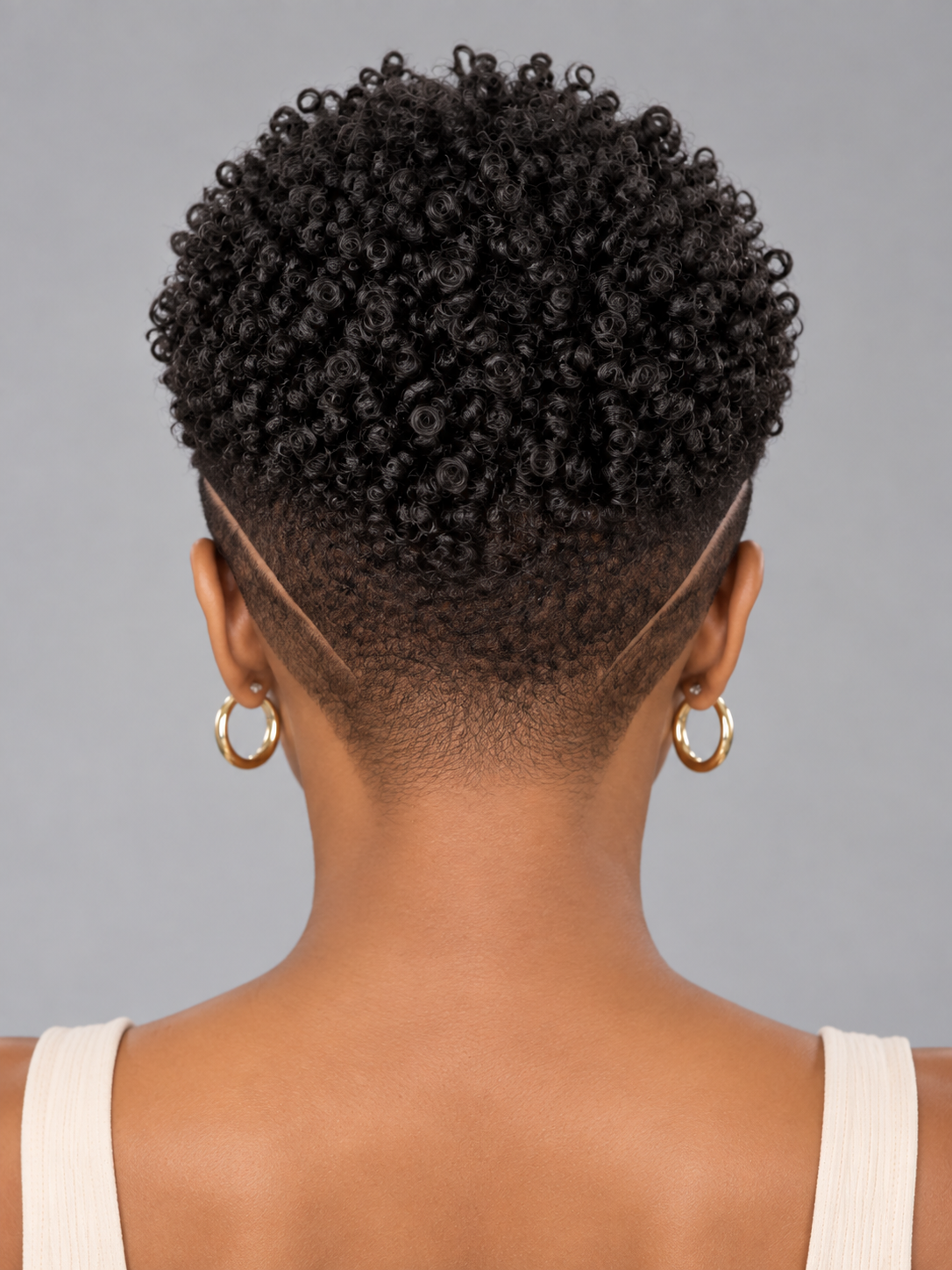 Back view of a person with a short, curly black hairstyle, wearing gold hoop earrings and a white ribbed tank top against a solid gray background.