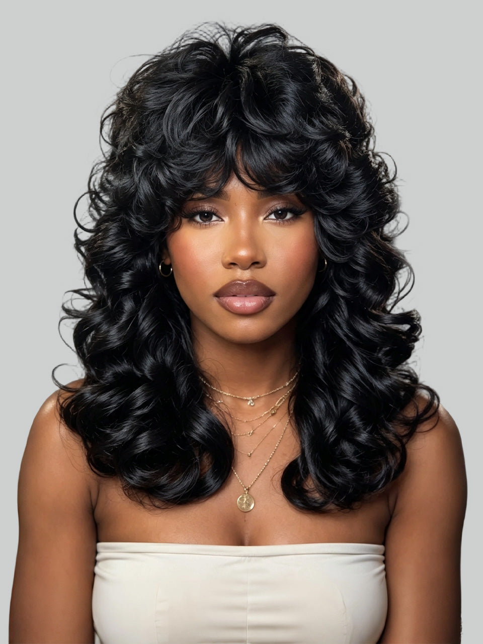 A woman with long, voluminous, black curly hair wearing a strapless cream top, photographed against a solid light gray background.