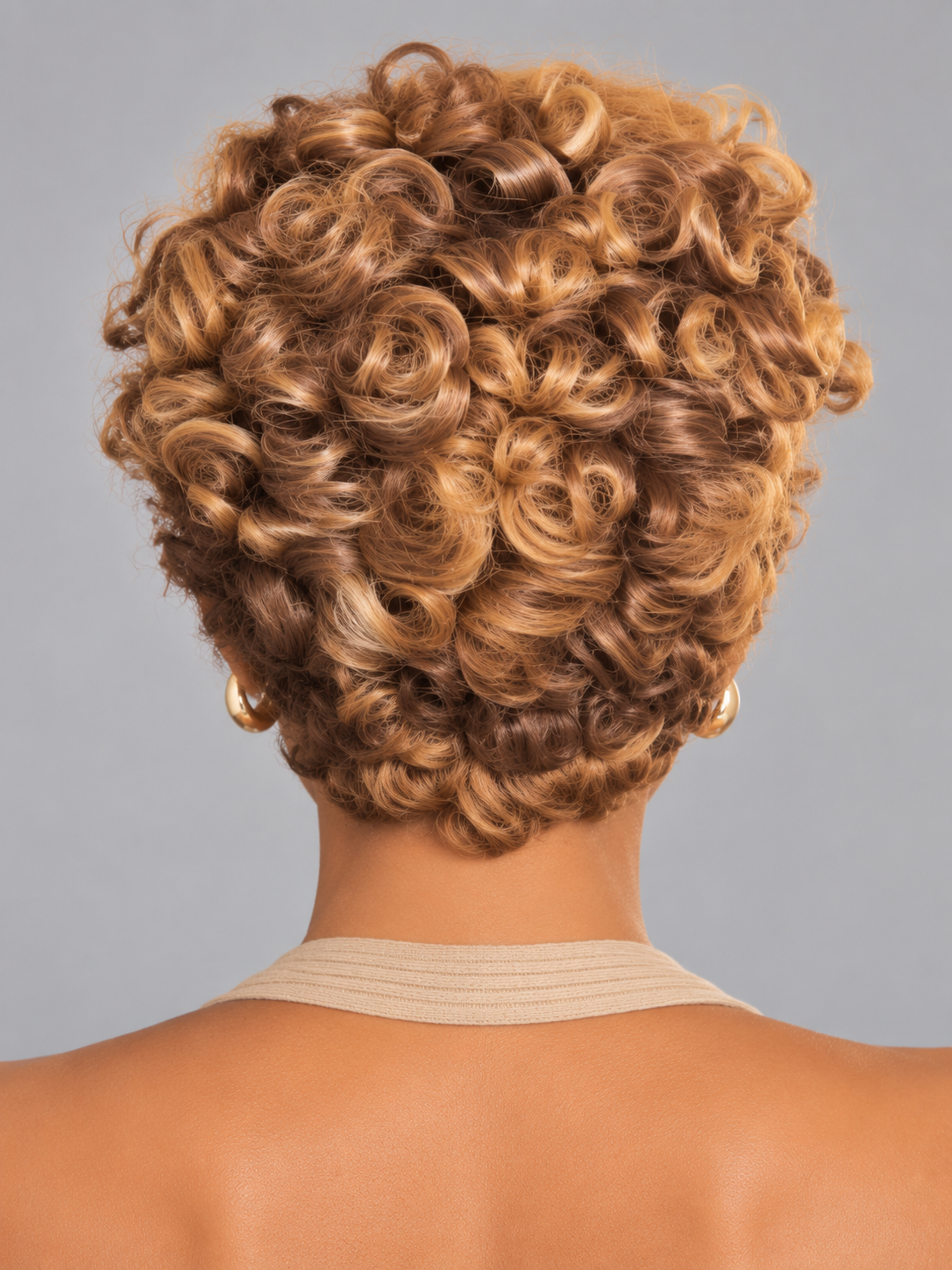 Back view of a person with a short, tightly curled hairstyle in warm golden-brown tones, wearing a beige ribbed halter top against a plain gray studio background.