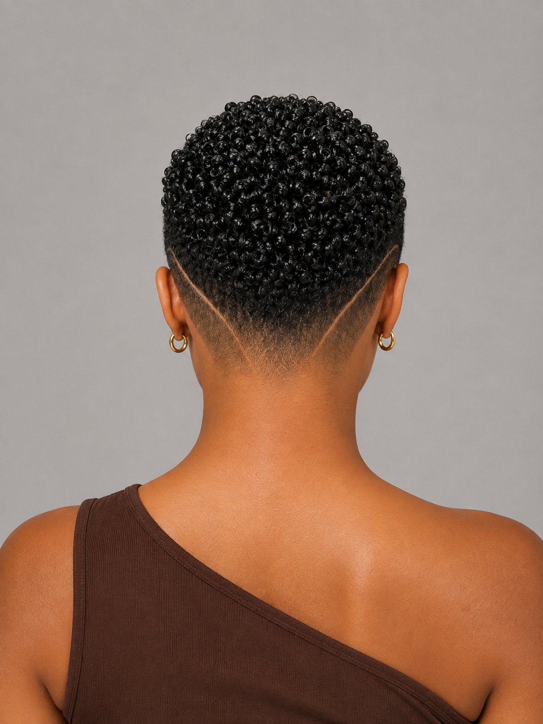 Back view of a person with a short, tightly curled black hairstyle and a side-swept fade, wearing small gold hoop earrings and a brown one-shoulder top against a plain gray background.