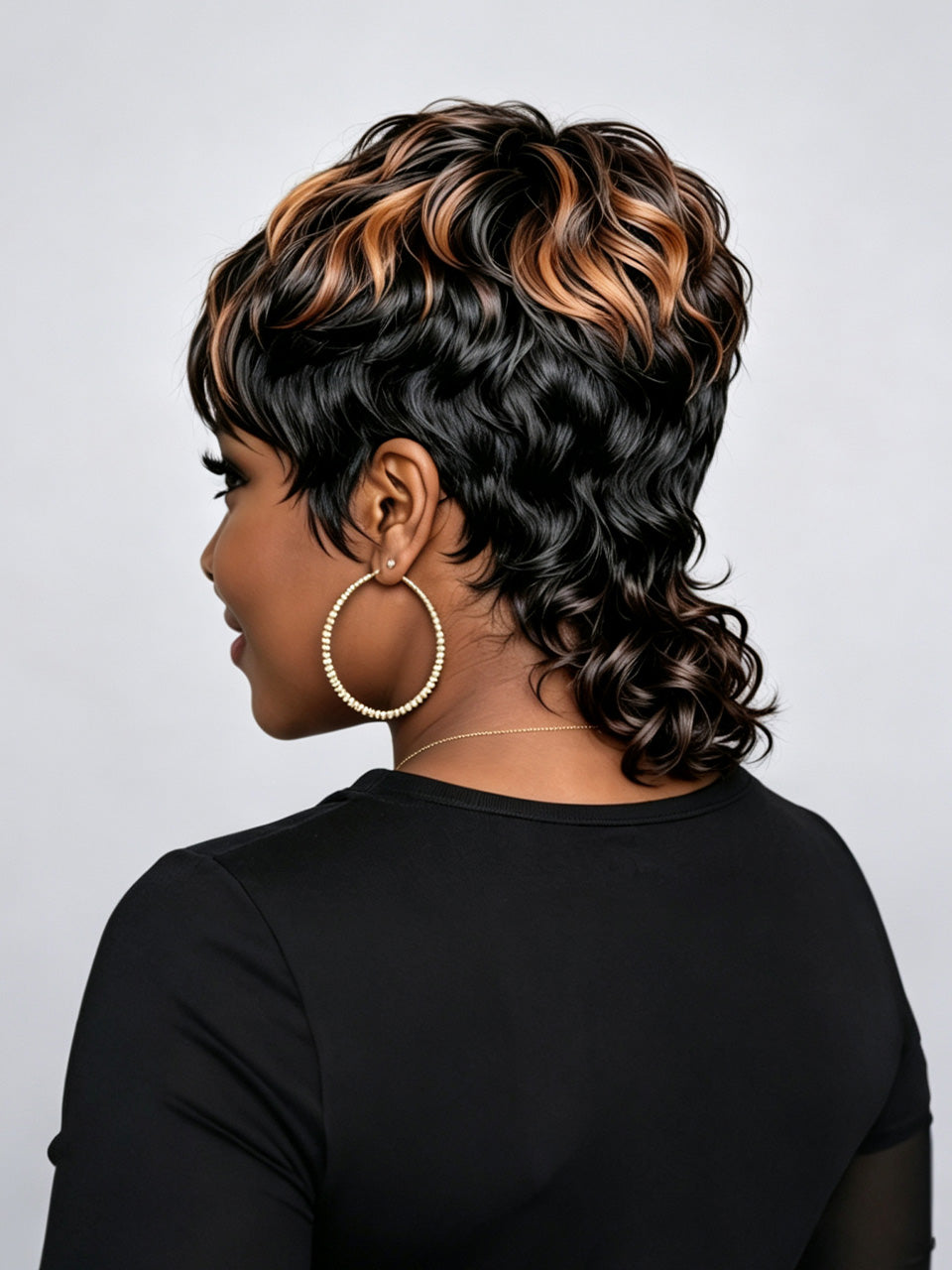 A woman shown from the back wearing a short, curly wig with black, brown, and warm blonde highlights, paired with large gold hoop earrings and a black top against a light gray background.