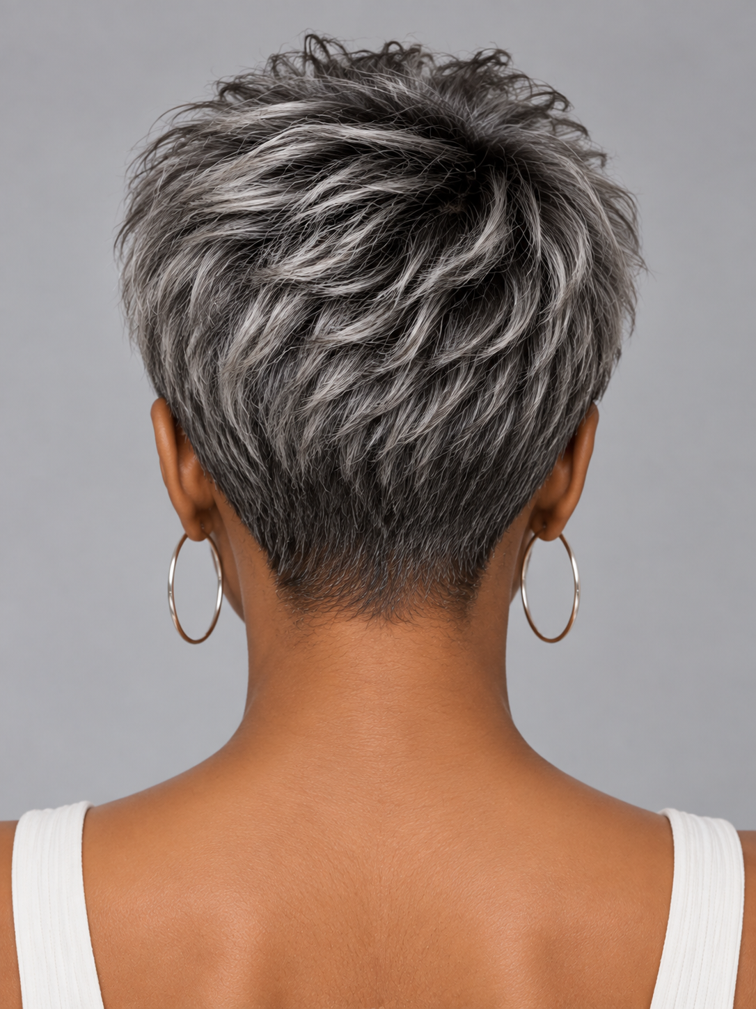 Back view of a person with a short, layered pixie cut featuring mixed silver-gray and dark brown hair, wearing large gold hoop earrings and a white tank top against a solid gray background.