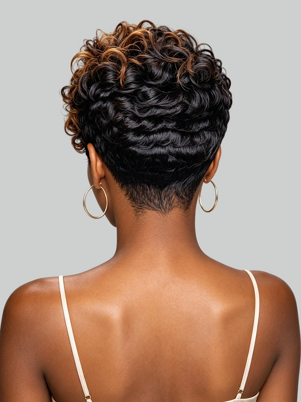Back view of a woman with a short, curly, textured wig in dark brown with warm caramel highlights, paired with gold hoop earrings and a white spaghetti strap top, set against a solid gray background.