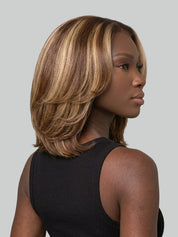 A woman shown from the back and side profile wearing a shoulder-length layered wig with warm brown and blonde highlights, against a solid light gray background.