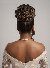 Back view of a woman with an intricate braided updo hairstyle, wearing a pearl necklace and an off-the-shoulder lace wedding dress against a neutral gray background.