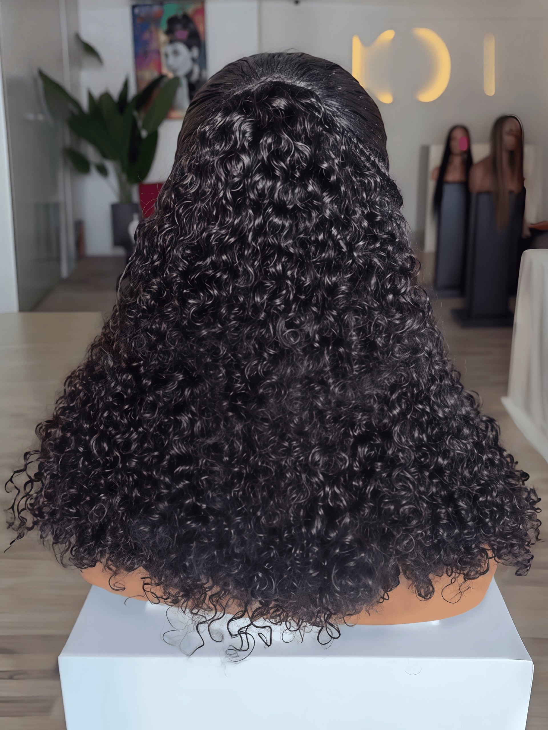 A black curly wig displayed on a mannequin head stand, viewed from the back in a salon-like interior with a blurred background.
