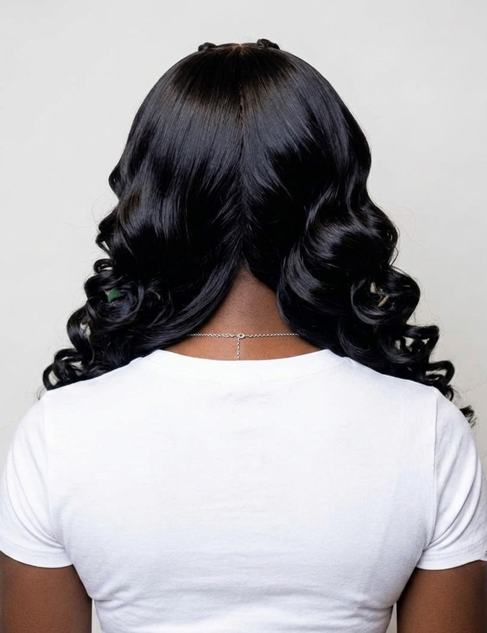 Back view of a person with long, glossy black wavy hair, wearing a plain white t-shirt against a light gray background.