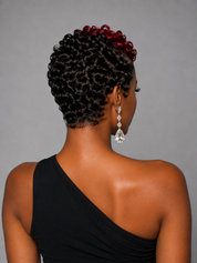 A woman shown from the back wearing a black one-shoulder dress, with her hair styled in tight curls and a dark red top section. She is wearing dangling clear teardrop earrings against a plain gray background.