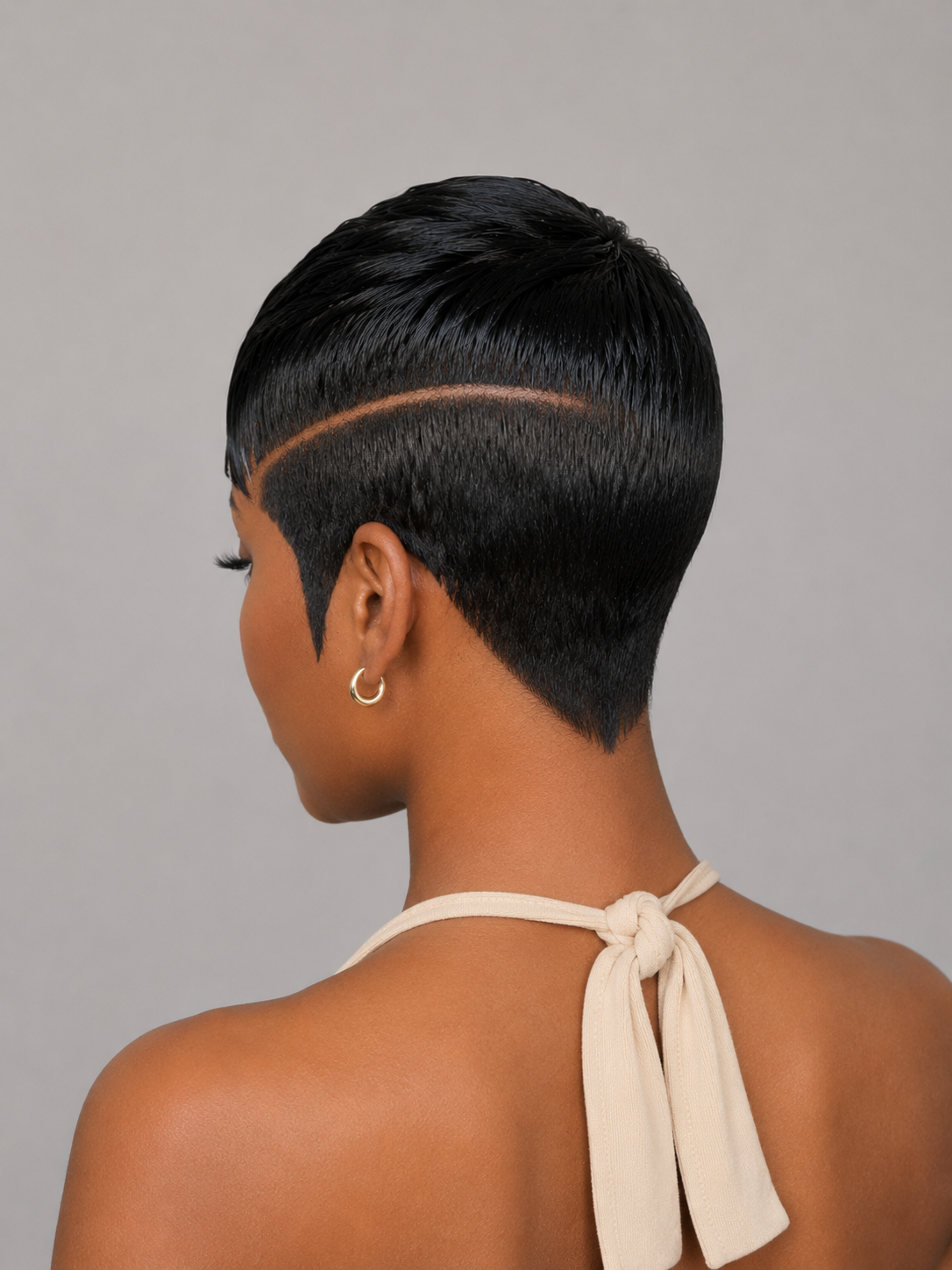 Rear view of a person with a short, dark black textured hairstyle featuring a precise side part, wearing a cream halter top against a solid gray background.