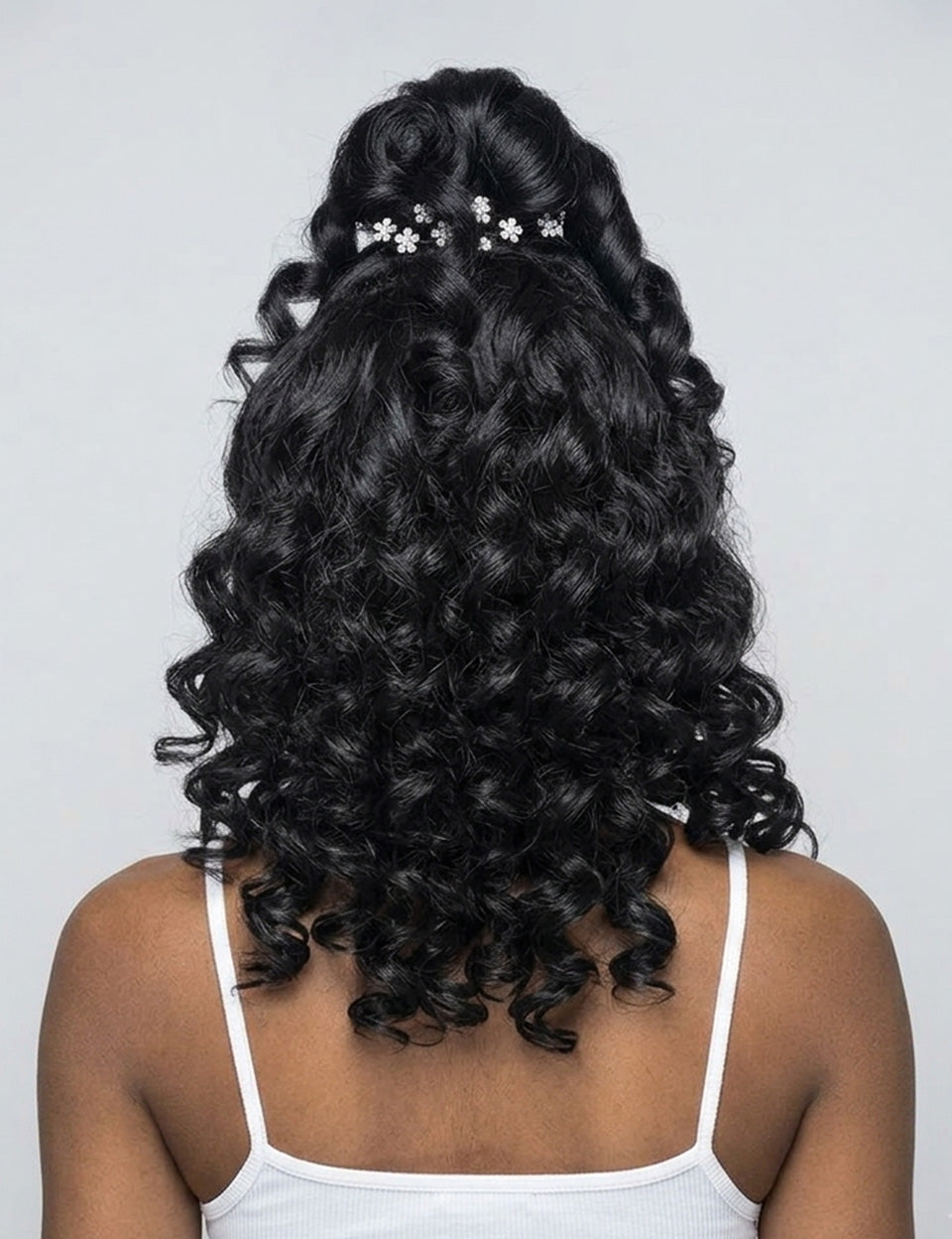 Back view of a person with long, dark, curly hair styled in a half-updo with small white floral hair clips, wearing a white spaghetti-strap top against a light gray background.