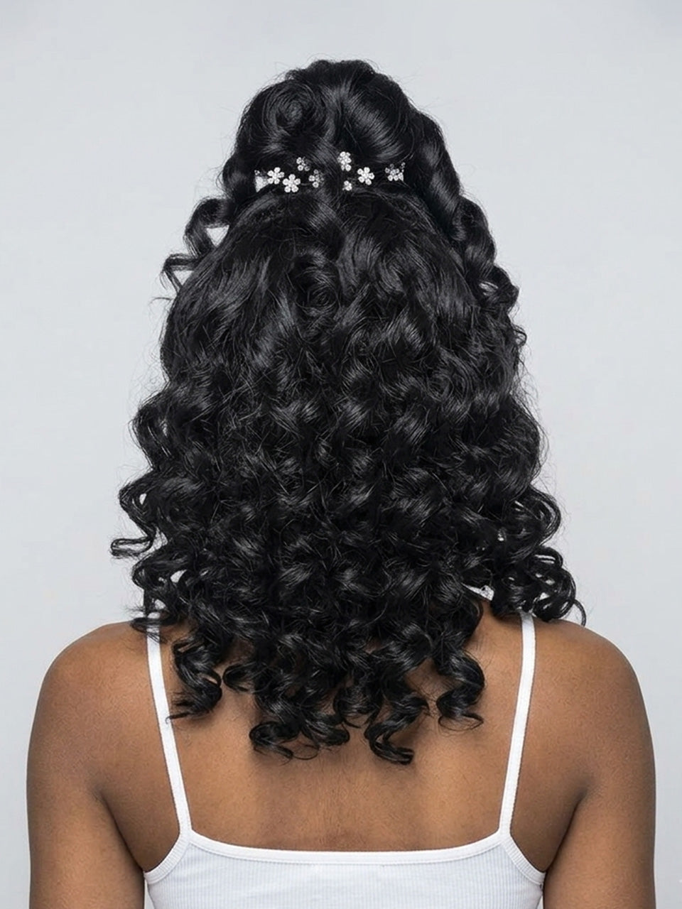 Back view of a person with long, dark, curly hair styled in a half-updo with small white floral hair clips, wearing a white spaghetti-strap top against a light gray background.