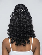 Back view of a person with long, dark, curly hair styled in a half-updo with small white floral hair clips, wearing a white spaghetti-strap top against a light gray background.