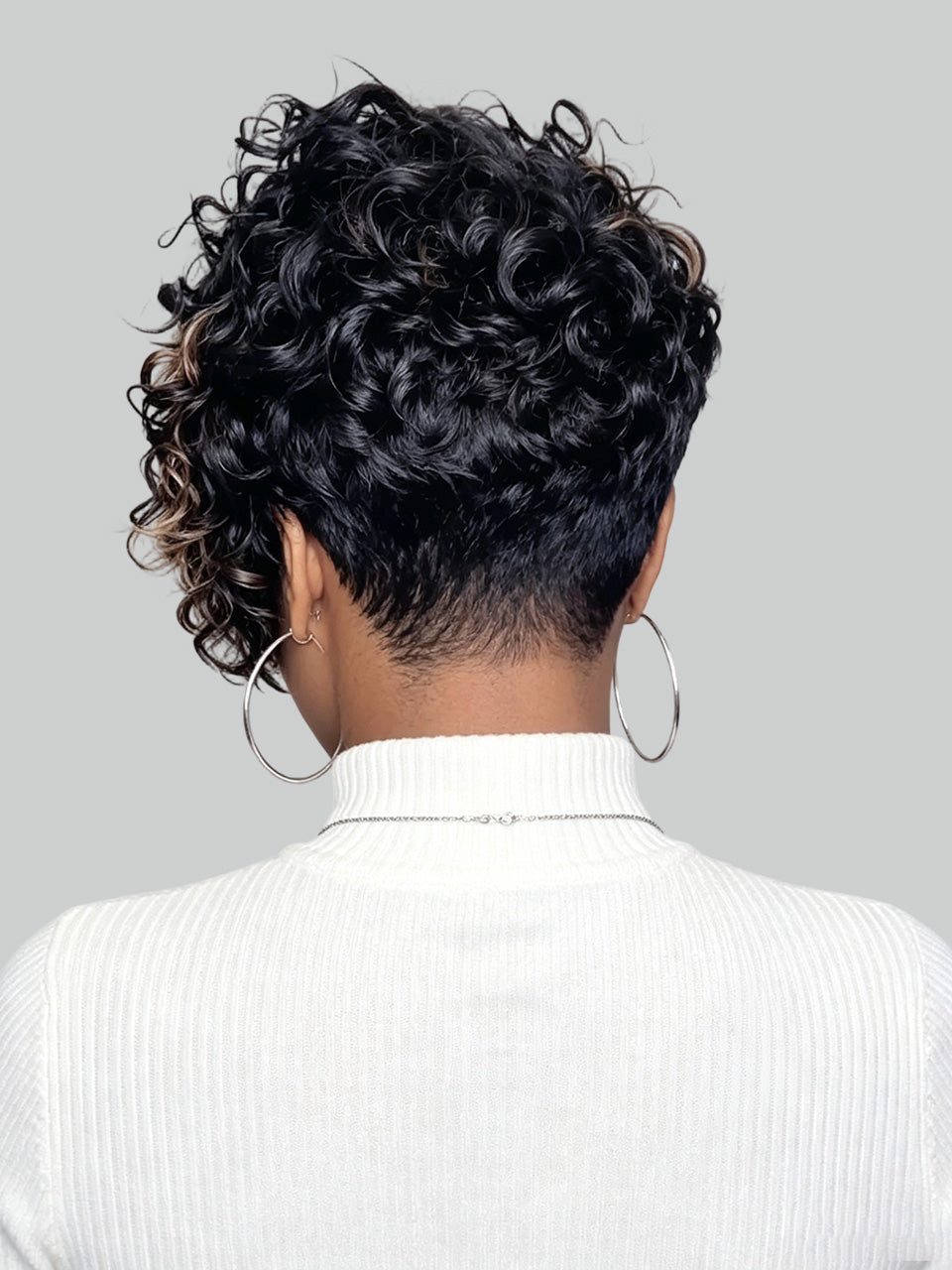 Back view of a person with a short, curly black wig or hairpiece, wearing a white ribbed turtleneck sweater and large silver hoop earrings against a light gray background.