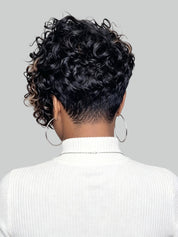 Back view of a person with a short, curly black wig or hairpiece, wearing a white ribbed turtleneck sweater and large silver hoop earrings against a light gray background.