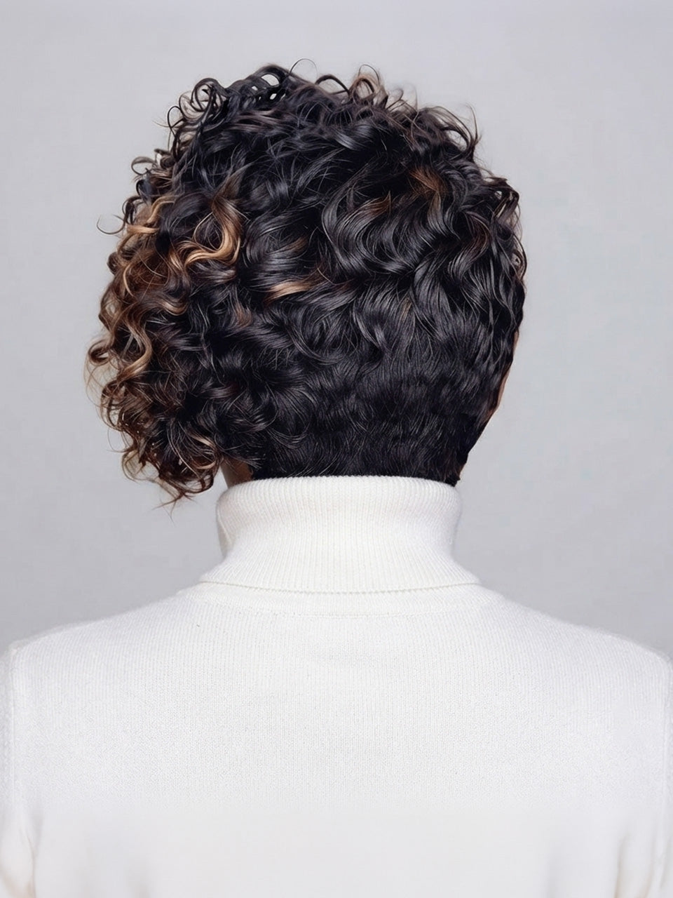 Back view of a person with short, curly dark hair featuring warm caramel-brown highlights, wearing a white turtleneck sweater against a light gray background.