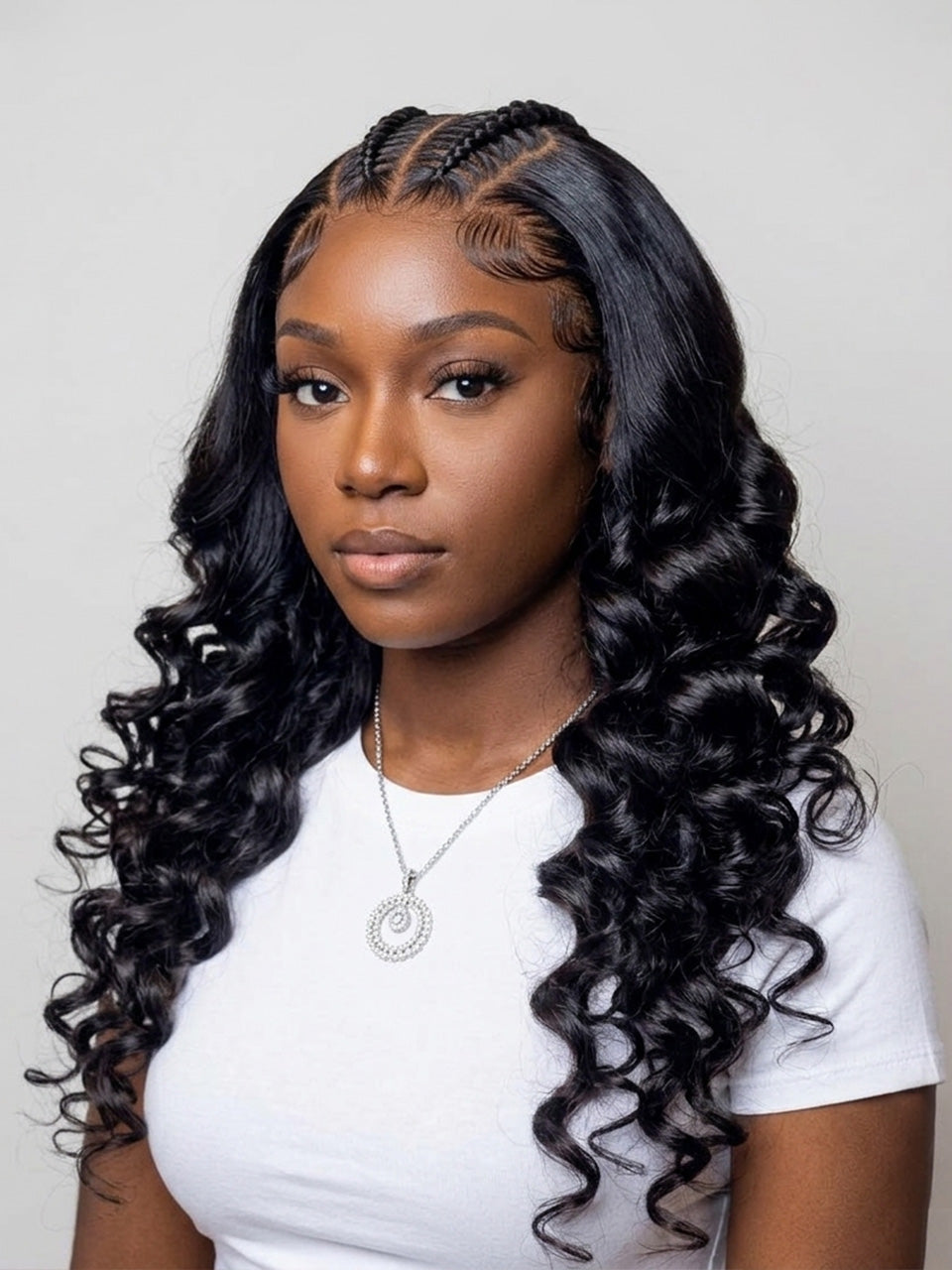 A woman with long, glossy, black curly wig with deep wave pattern, styled with braided top hair, wearing a white t-shirt and silver necklace against a light gray background.
