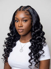 A woman with long, glossy, black curly wig with deep wave pattern, styled with braided top hair, wearing a white t-shirt and silver necklace against a light gray background.