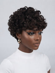 A woman wearing a short, dark brown curly wig with a lace front, styled with dramatic winged eyeliner and glittery eye makeup, wearing a white ribbed t-shirt against a light gray background.