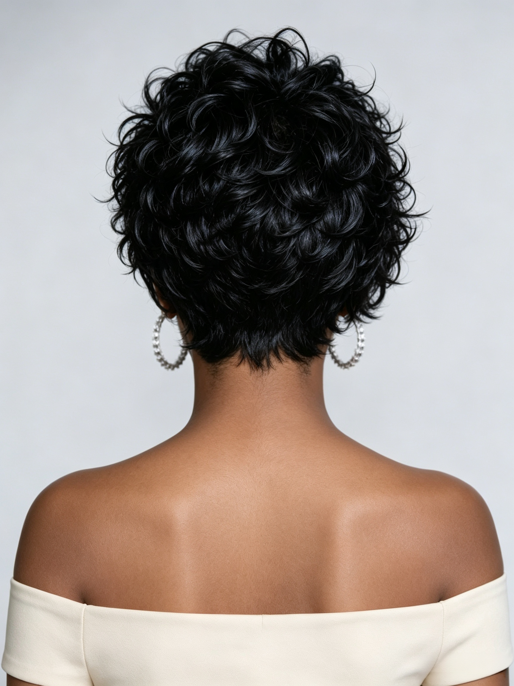 Rear view of a person with a short, curly black wig or hairpiece, wearing large pearl hoop earrings and an off-the-shoulder cream top against a light gray studio background.