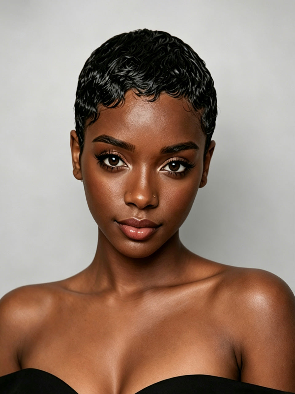 A close-up portrait of a woman with short, textured black hair, wearing a black strapless top against a light gray studio background.