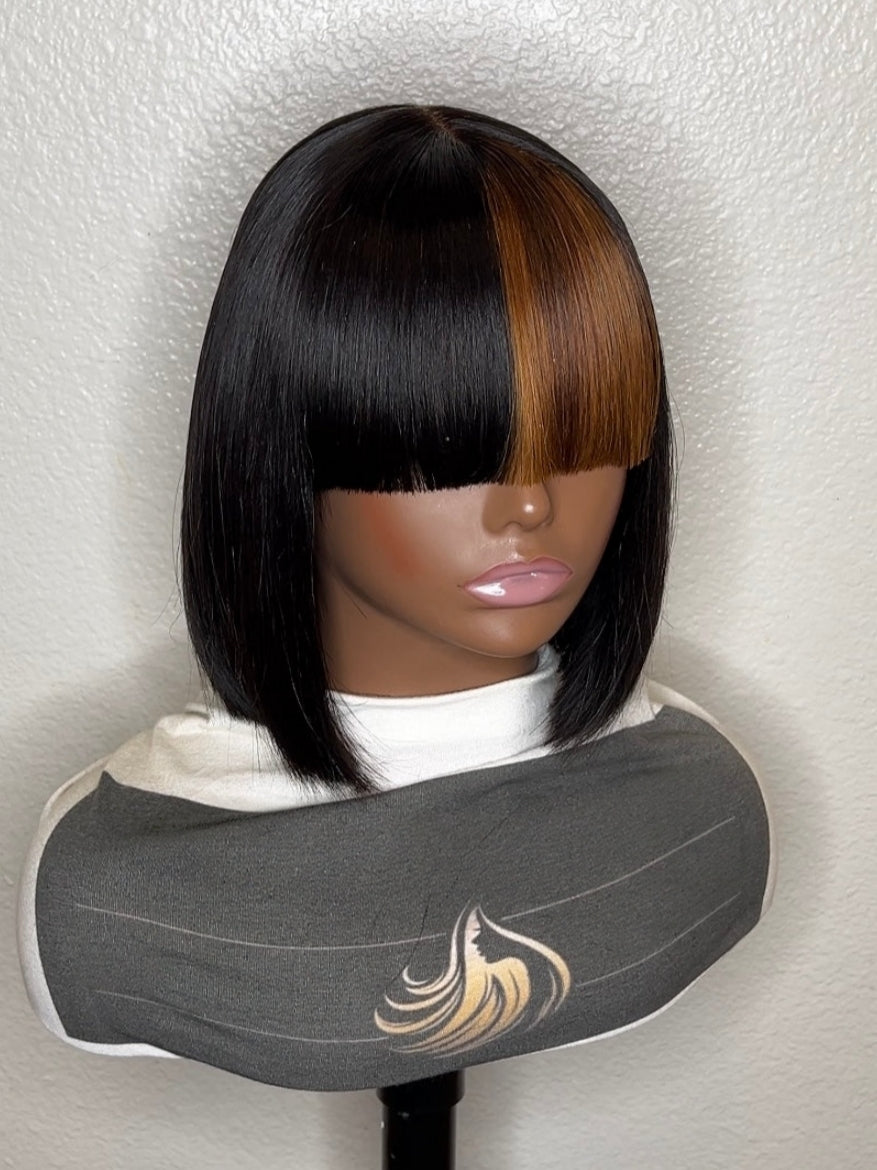 A short straight wig displayed on a mannequin head and neck form against a textured off-white wall. The wig has a dark black front section with full bangs, a center part, and warm medium brown highlights running down the right side.