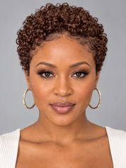 A woman with short, tightly curled brown hair wearing large gold hoop earrings and a white ribbed top, shown against a plain gray studio background.