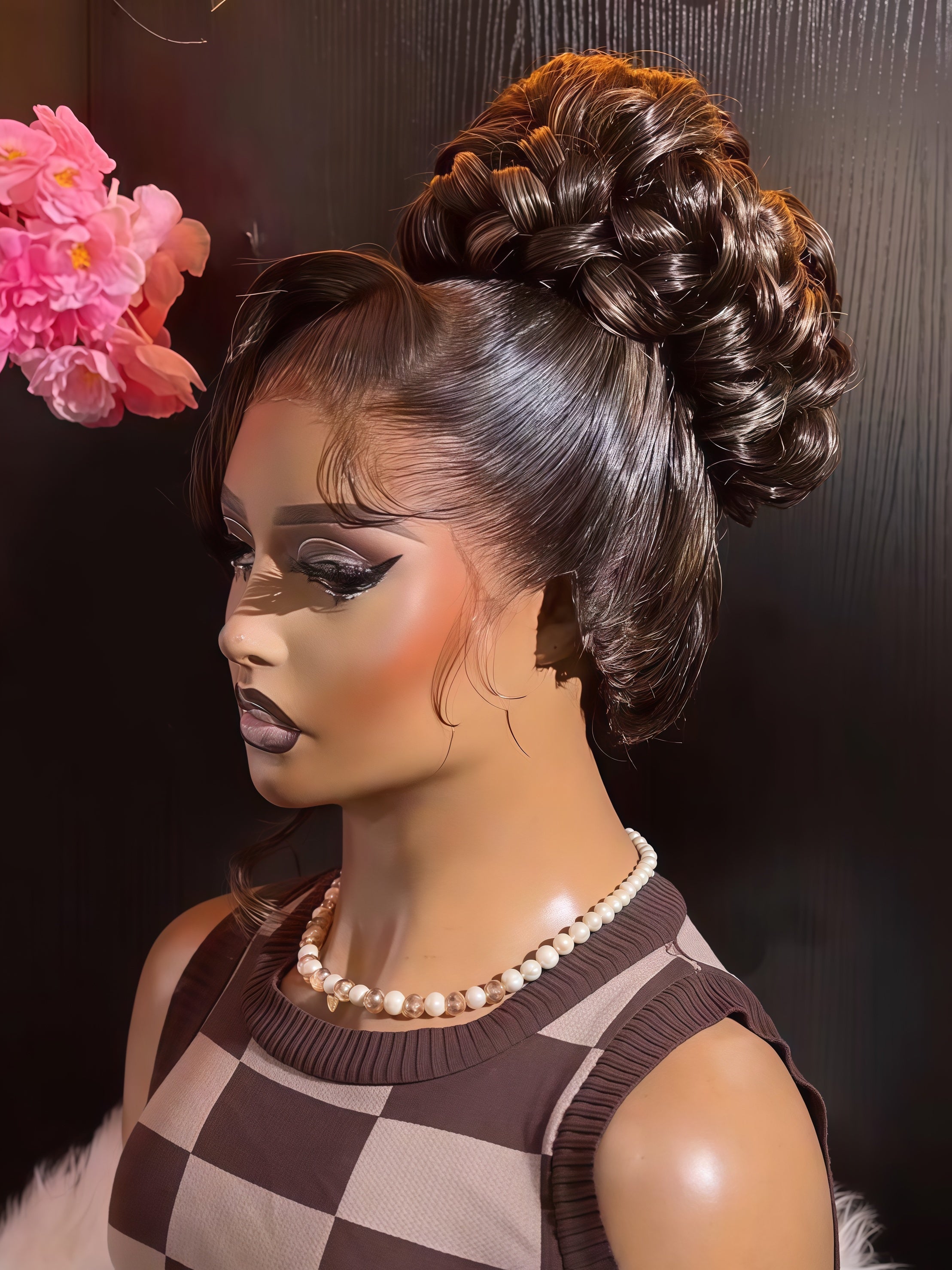 A close-up of a mannequin or doll with a voluminous dark brown updo hairstyle, wearing a brown and beige checkered sleeveless top and a pearl necklace against a dark background.