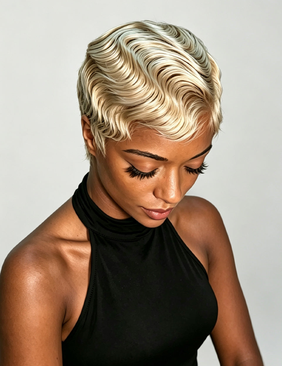 A woman wearing a short, wavy platinum blonde wig, looking down, dressed in a black halter top against a light gray background.