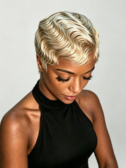A woman wearing a short, wavy platinum blonde wig, looking down, dressed in a black halter top against a light gray background.