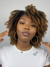 A woman with voluminous, curly brown hair wearing a white t-shirt, gold hoop earrings, and a gold chain necklace, posing against a light gray background.