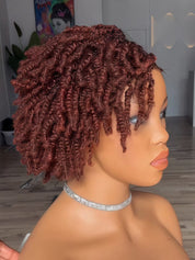 A short, curly wig displayed on a mannequin head in profile, featuring tight, glossy ringlets in a deep auburn reddish-brown color. The mannequin is shown indoors with a blurred background of a room.
