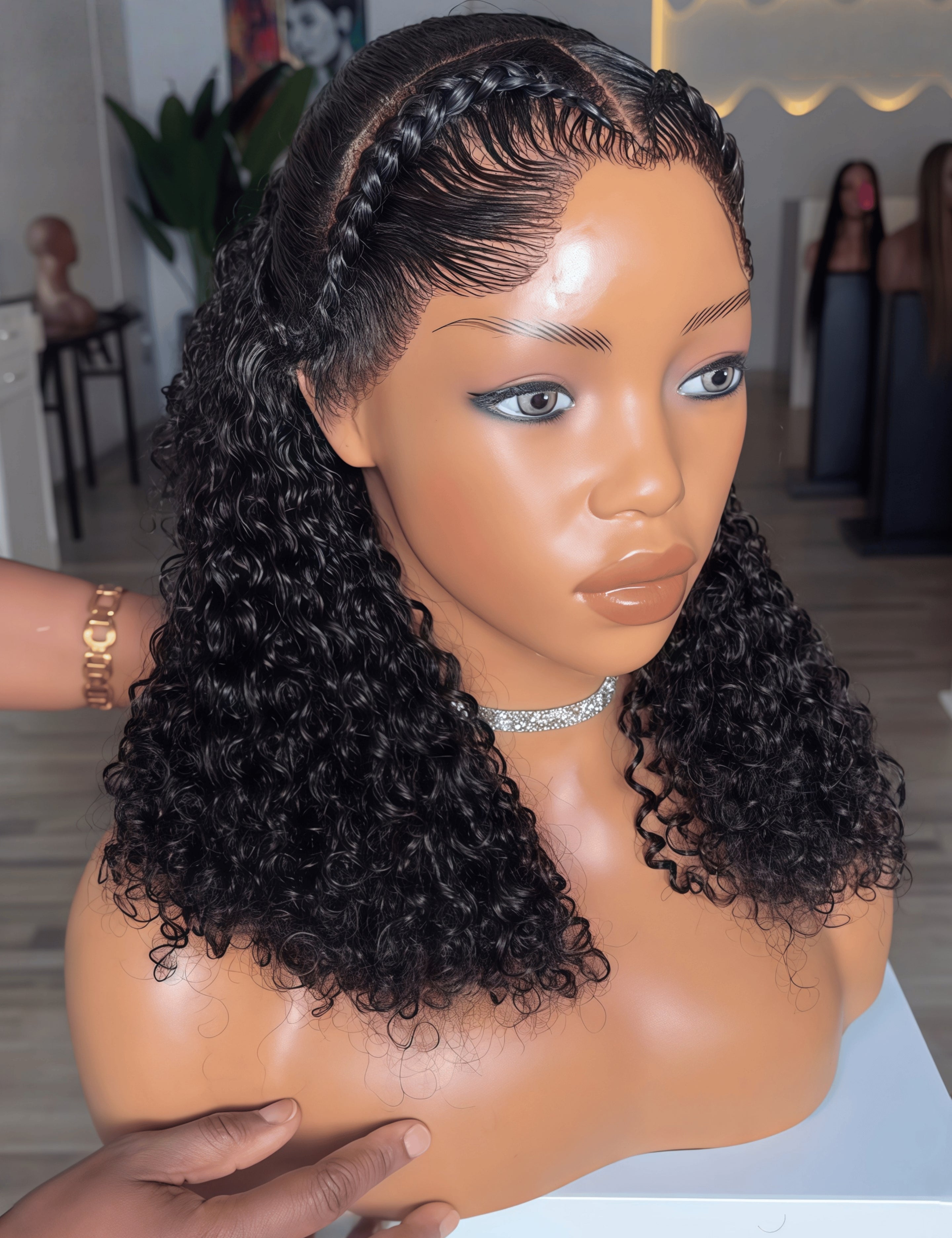 A mannequin bust wearing a dark curly wig with deep wave curls and braided hair along the crown, photographed indoors with a blurred boutique background.