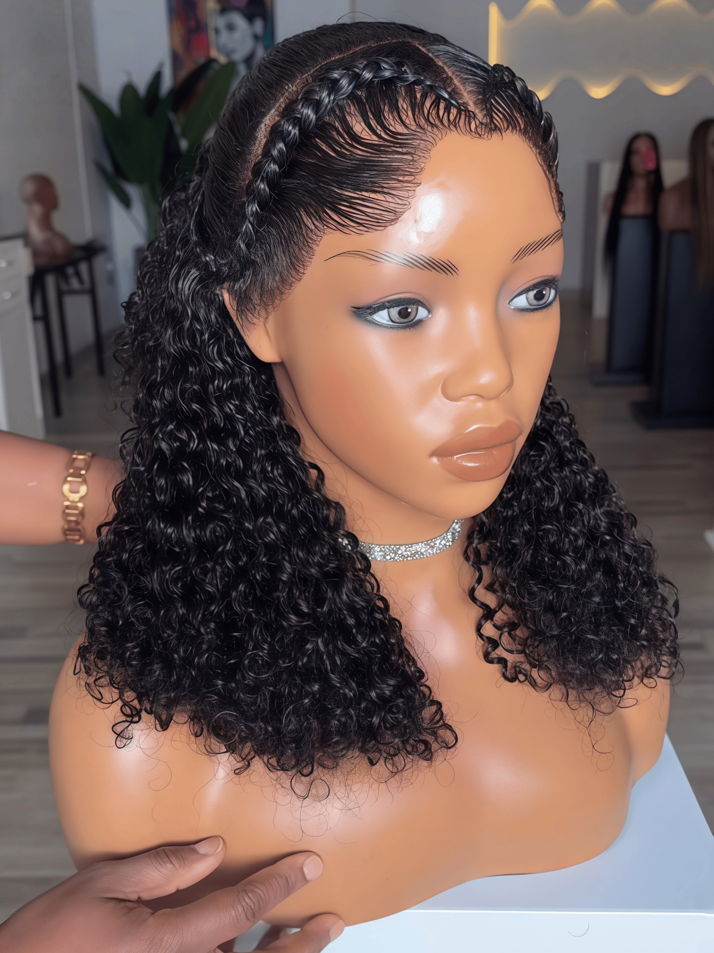A mannequin bust wearing a dark curly wig with deep wave curls and braided hair along the crown, photographed indoors with a blurred boutique background.