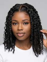 A woman with long, dark, curly hair wearing a white t-shirt, posing against a solid gray background. She is touching one side of her hair with her hand.