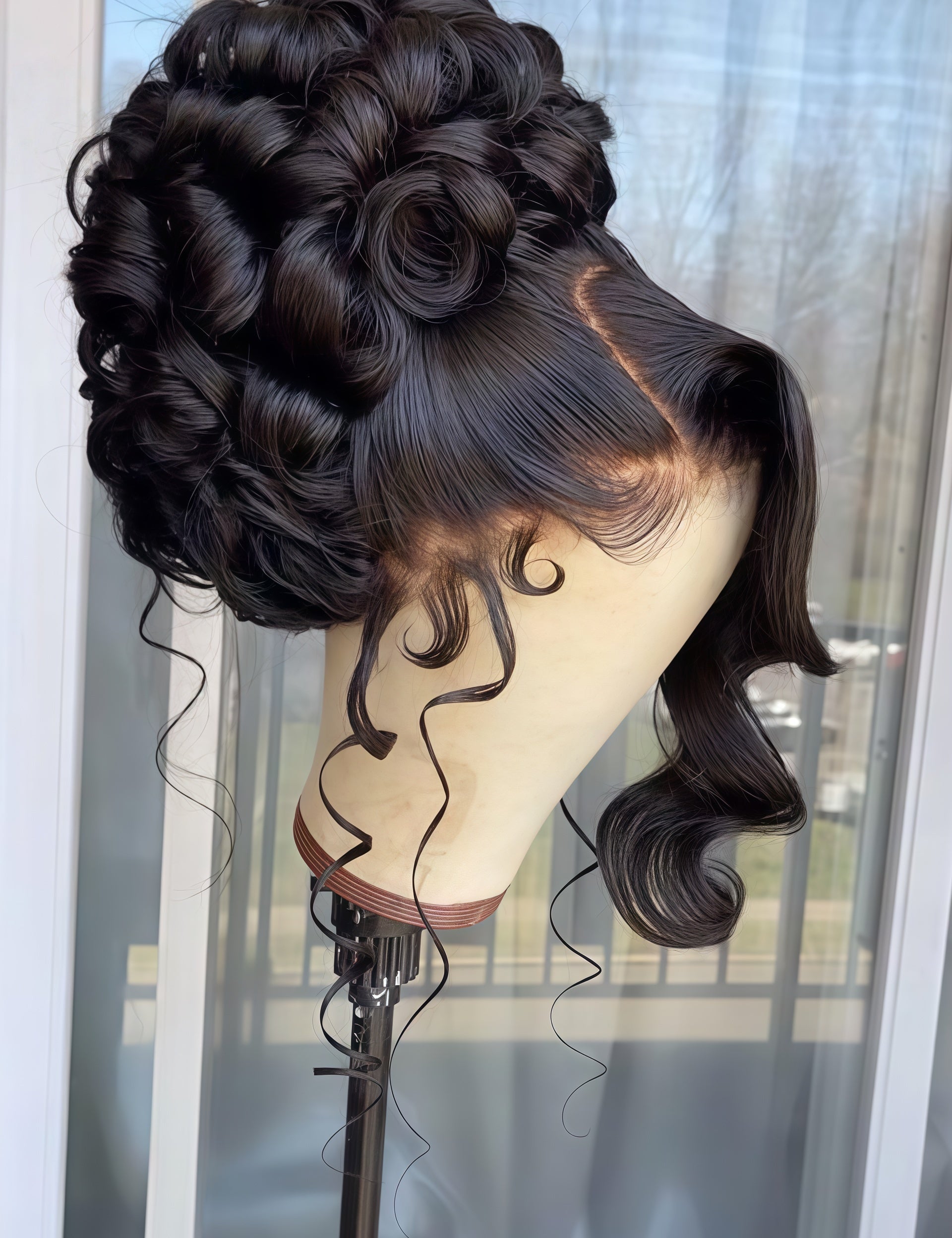 A dark brown to black wavy lace front wig styled in an elegant updo with curls, displayed on a mannequin head stand in front of a window.