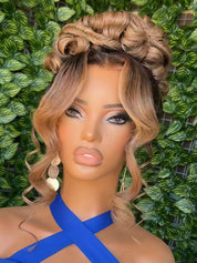 A close-up of a mannequin or doll with a blonde and brown ombre wig styled in an updo with side-swept curls, wearing a bright blue top against a green leafy background.
