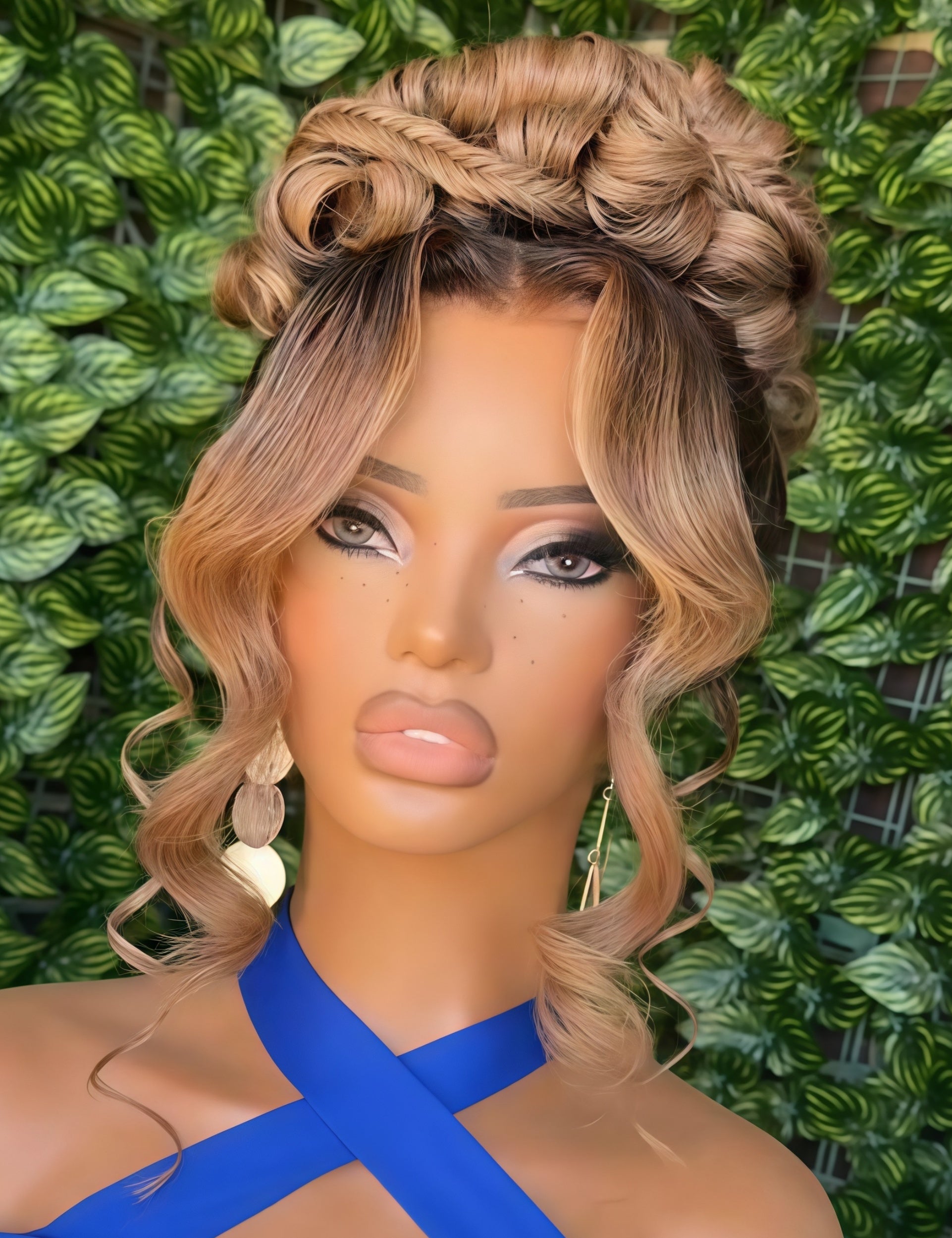 A close-up of a mannequin or doll with a blonde and brown ombre wig styled in an updo with side-swept curls, wearing a bright blue top against a green leafy background.