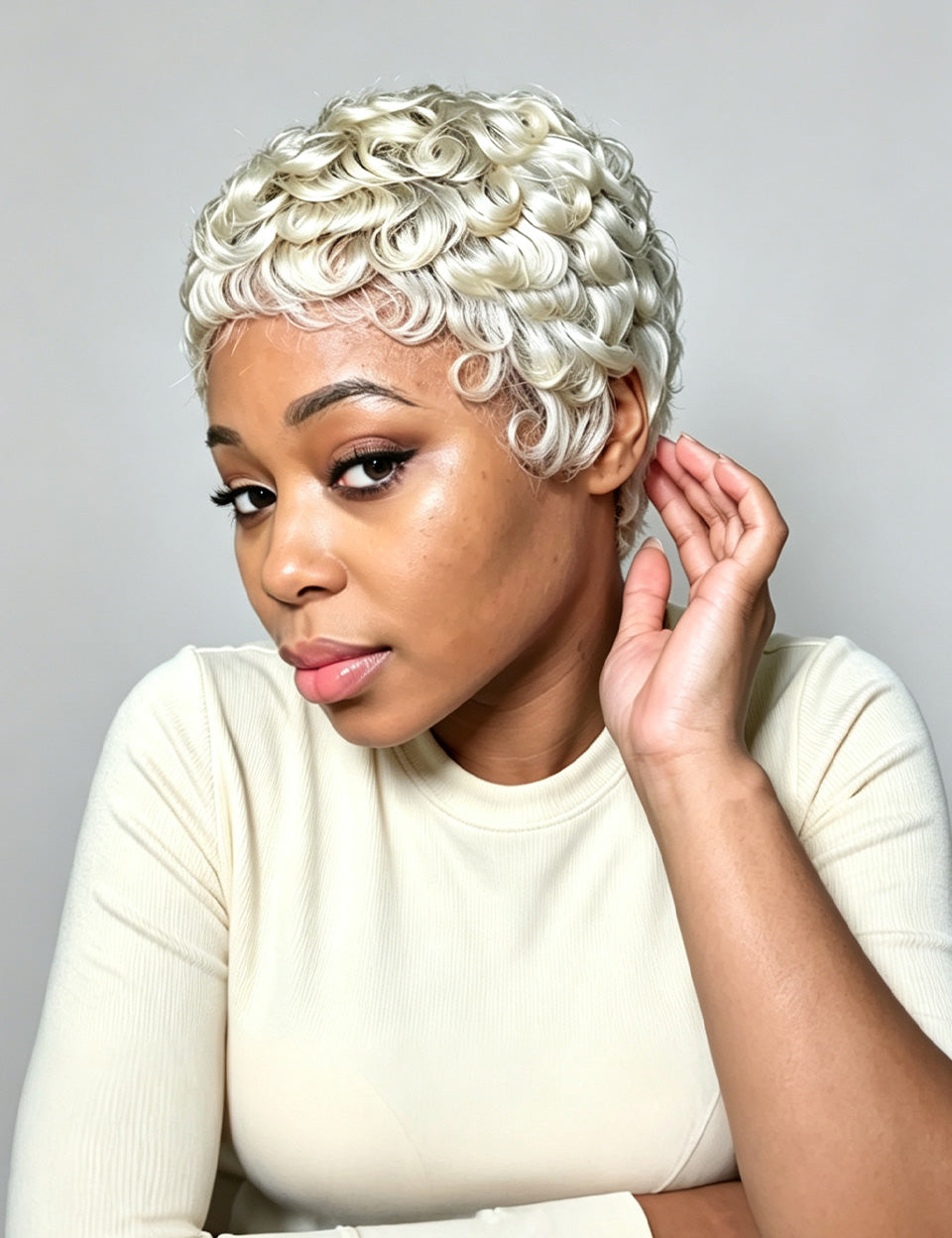 A woman with a short, curly platinum blonde wig wearing a cream ribbed long-sleeve top, posing against a neutral gray background.