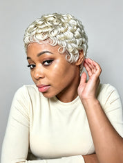 A woman with a short, curly platinum blonde wig wearing a cream ribbed long-sleeve top, posing against a neutral gray background.